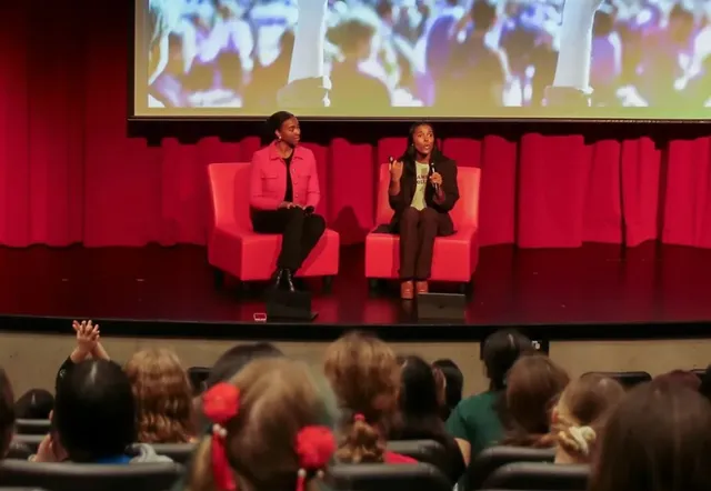 Nia and Justice in conversation on a red-lit stage in front of an auditorium of girls