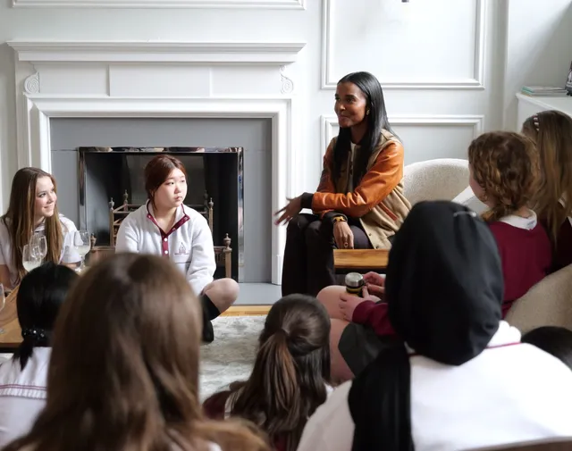 A Sisters founder in an orange varsity jacket sitting with a circle of schoolgirls in a panelled room