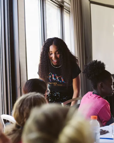 Nia Faith in a “Together We Move” shirt leaning in to talk with girls at a workshop table